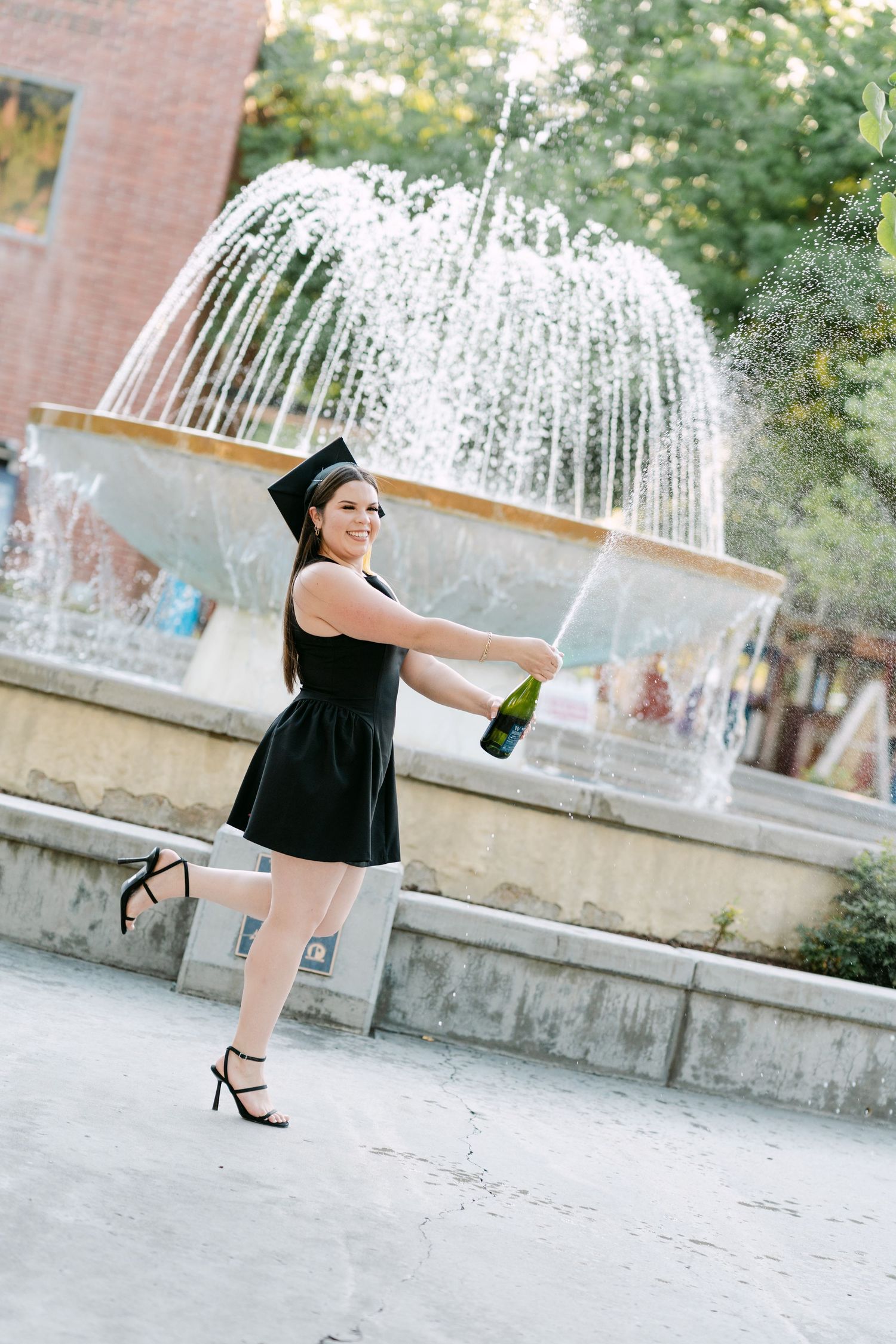 A graduate in a black dress and cap joyfully sprays champagne by a fountain outdoors.