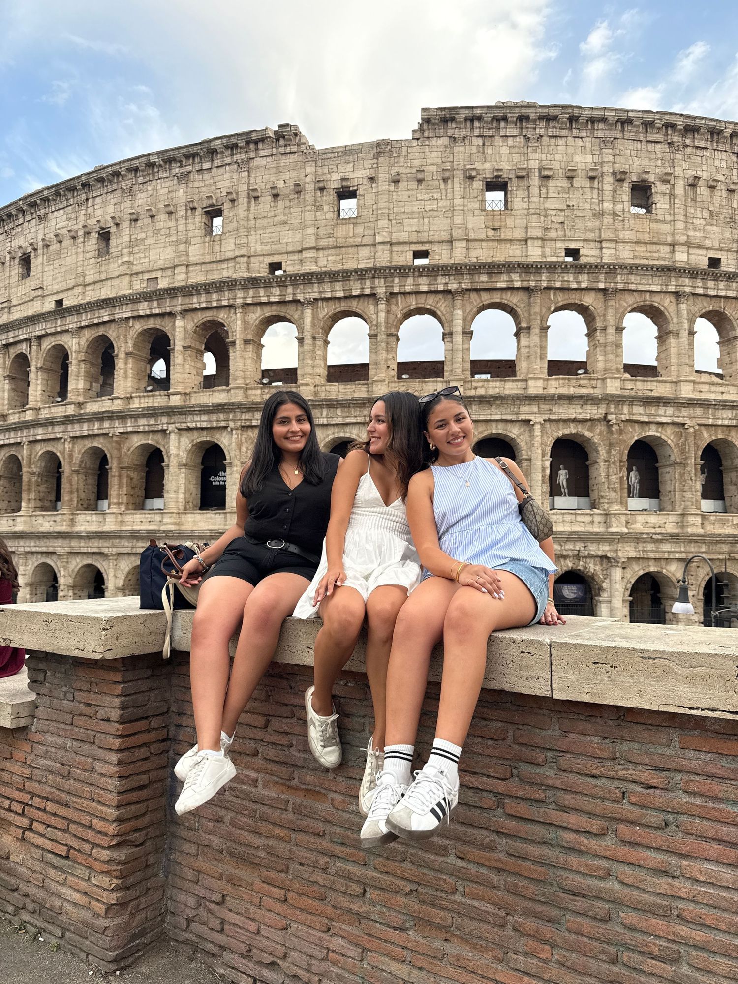 Three young women sitting on a wall in front of the iconic Colosseum in Rome, Italy on a sunny day.