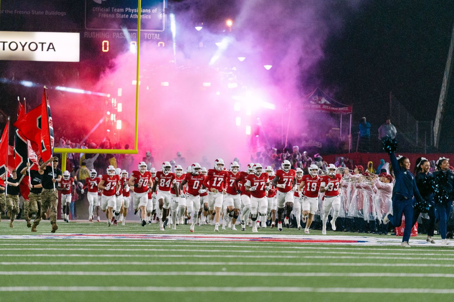 Football players run onto field through pink smoke and pyrotechnics during dramatic nighttime stadium entrance.
