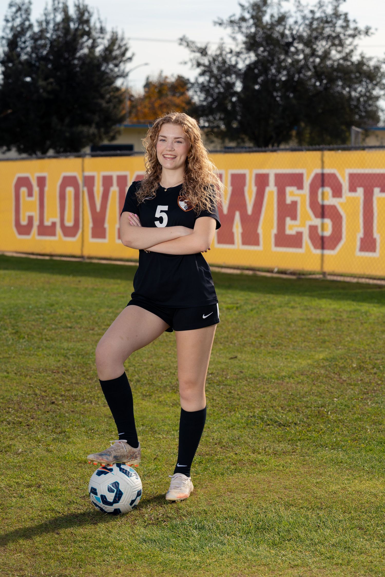 Soccer player in black uniform poses with ball on field with yellow 'CLOVIS WEST' sign in background.