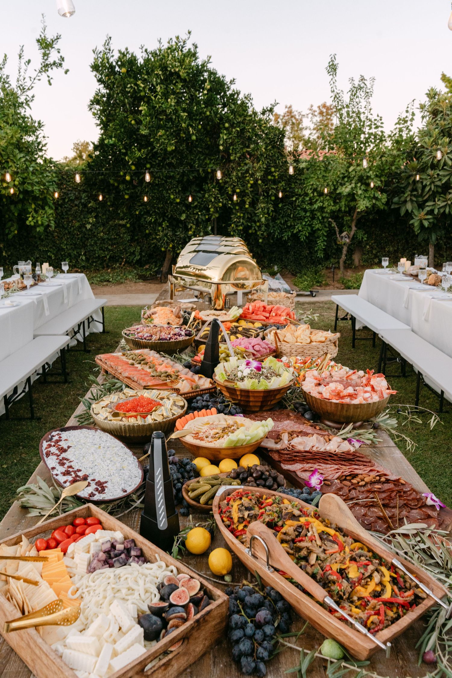 A rustic outdoor charcuterie board display featuring meats, cheeses, fruits, and snacks arranged on wooden platters at sunset.
