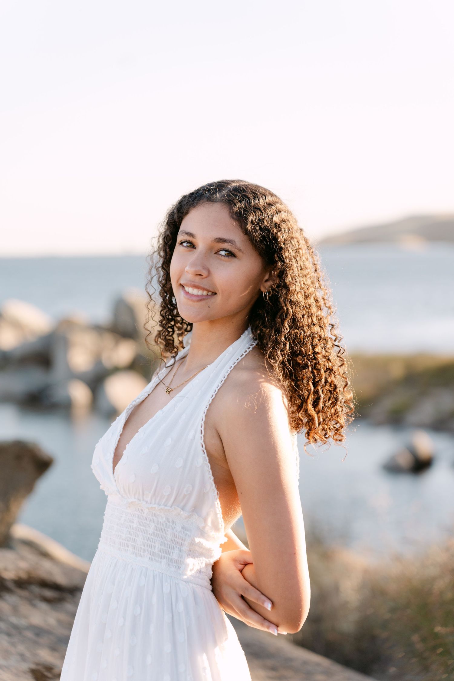 Young woman with curly hair in white halter dress smiling outdoors near rocky seaside at golden hour.
