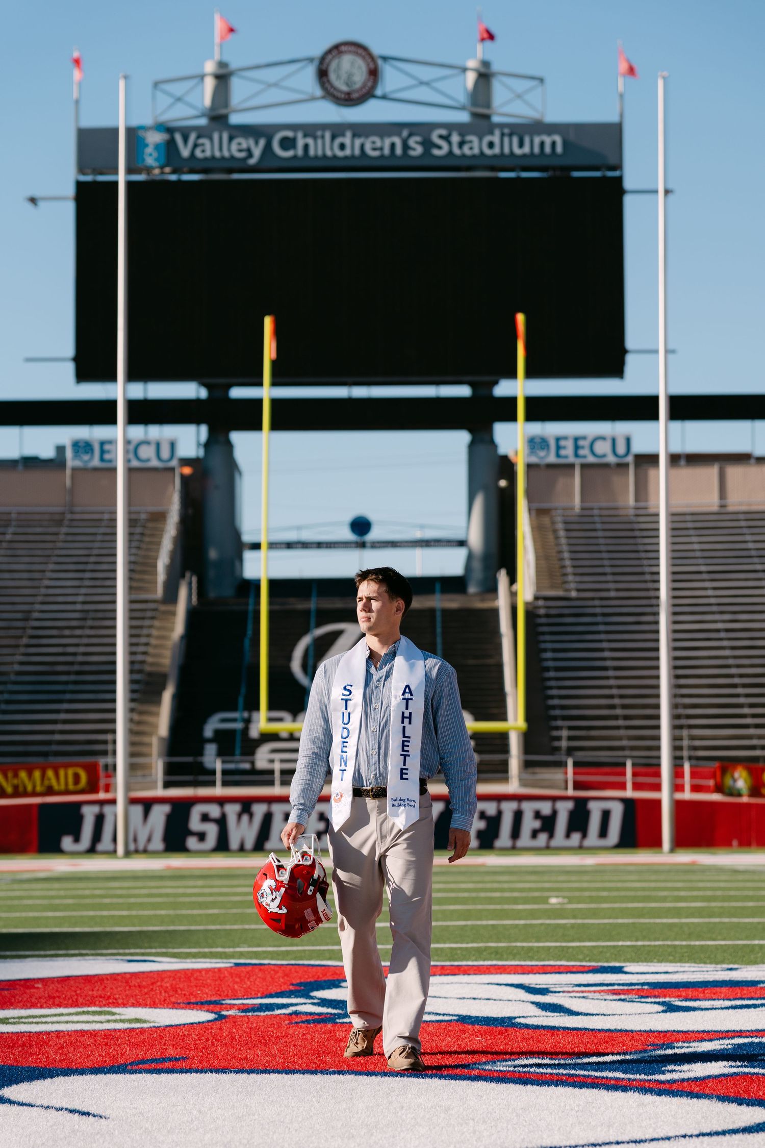 Graduate in stole walks on Jim Sweeney Field at Valley Children's Stadium, holding a red helmet.
