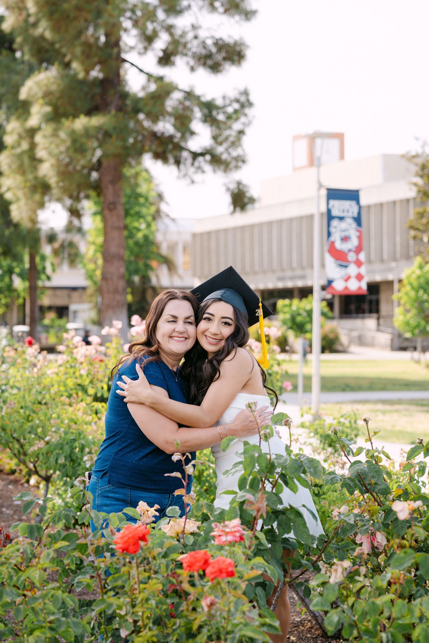 Two women hugging in a rose garden, one wearing a graduation cap and white dress, celebrating a college graduation outdoors.