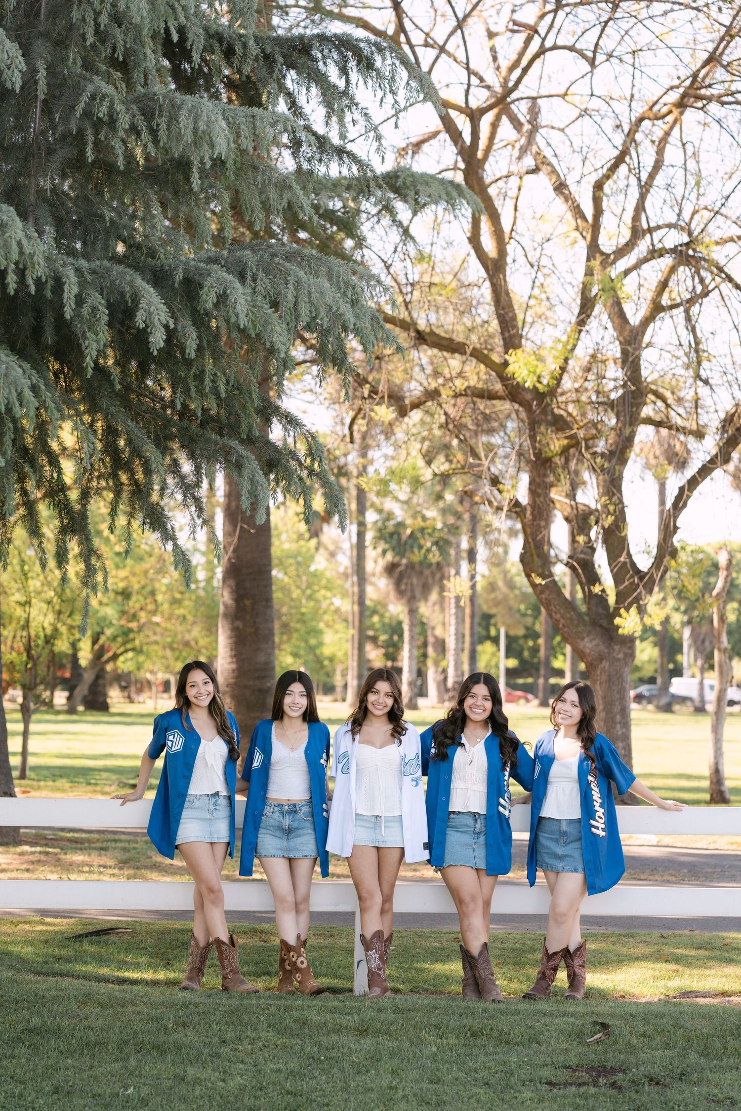 Five young women in blue jackets and denim shorts walk together in a sunny park with tall trees.