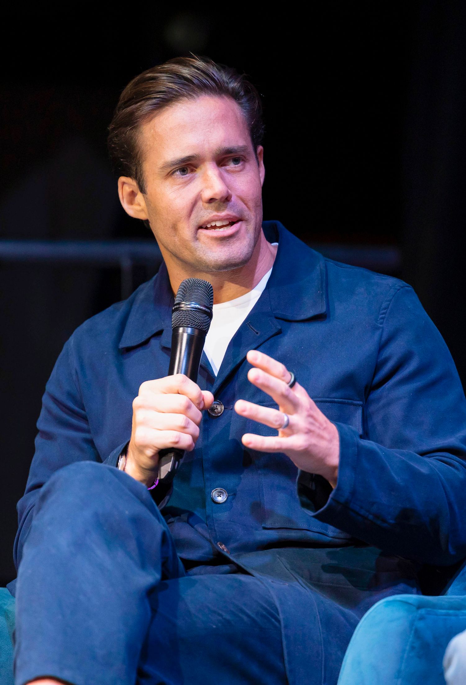 Speaker in blue jacket gesturing while talking into microphone on dark stage. London Event Photographer