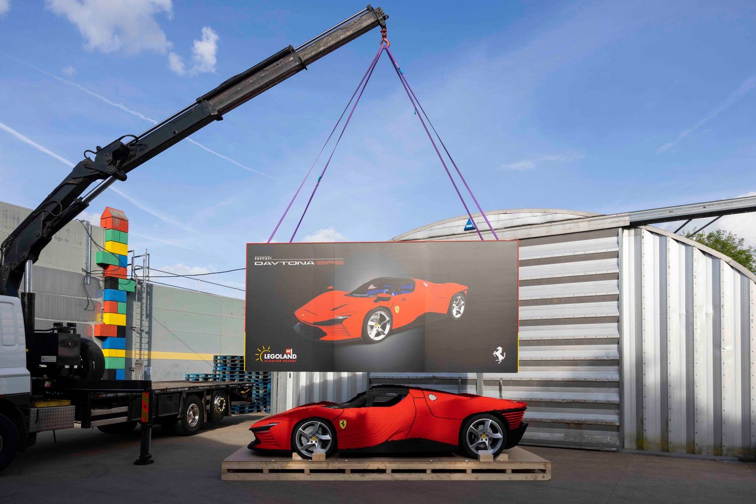 Red sports car parked next to a large promotional banner being lifted by a crane against a blue sky. London Event Photographer