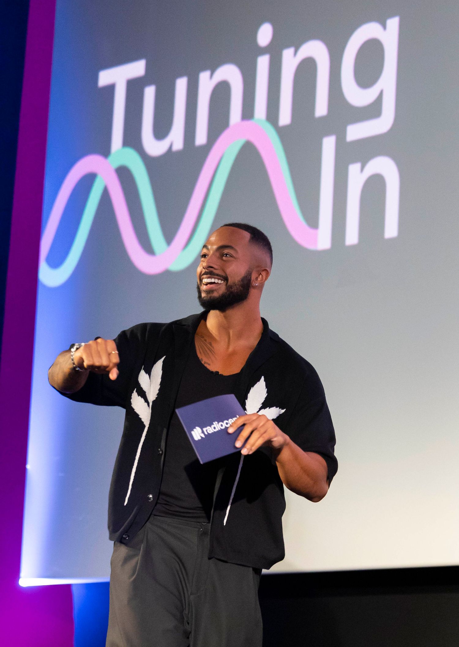 Presenter in black outfit speaking on stage with 'Tuning In' text and colorful wave graphic displayed at a london conference. London Event P