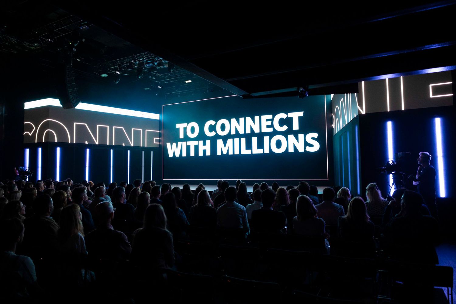 Large audience gathered in front of illuminated stage with text 'TO CONNECT WITH MILLIONS' displayed on screen. London Event Photographer