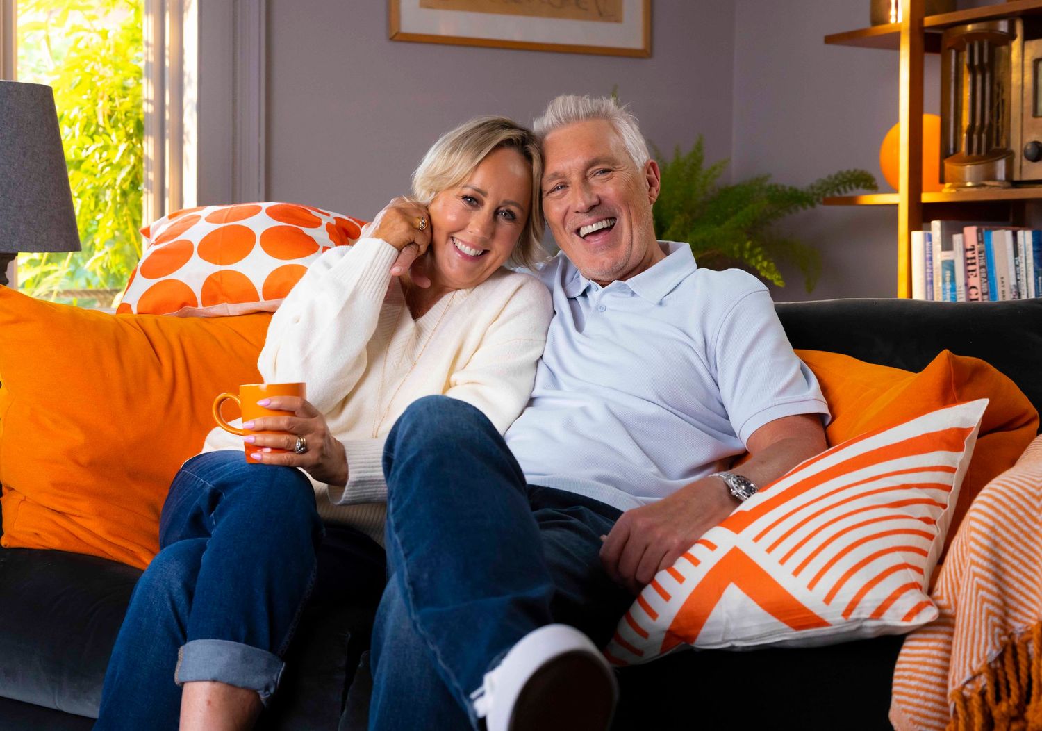Couple relaxing together on couch with orange pillows in sunlit living room. London Event Photographer