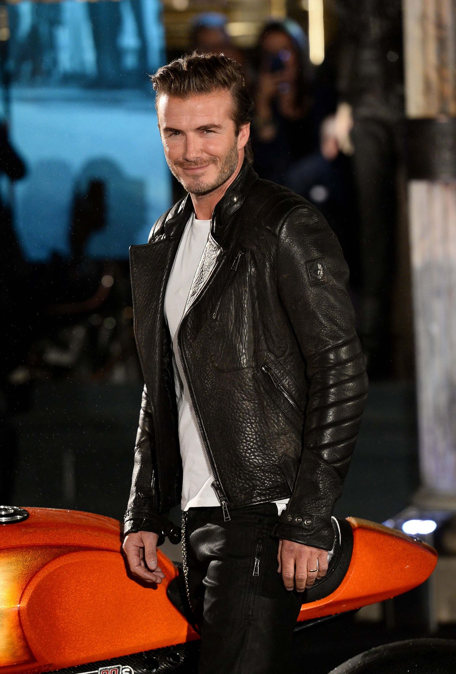 Person in leather jacket posing with orange motorcycle at night. London Event Photographer