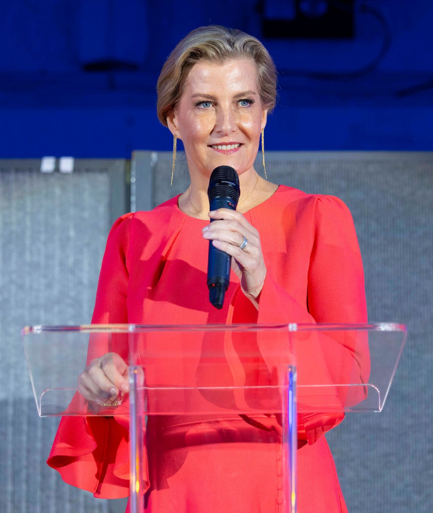 Speaker at podium with red outfit presenting on stage with transparent lectern. London Event Photographer