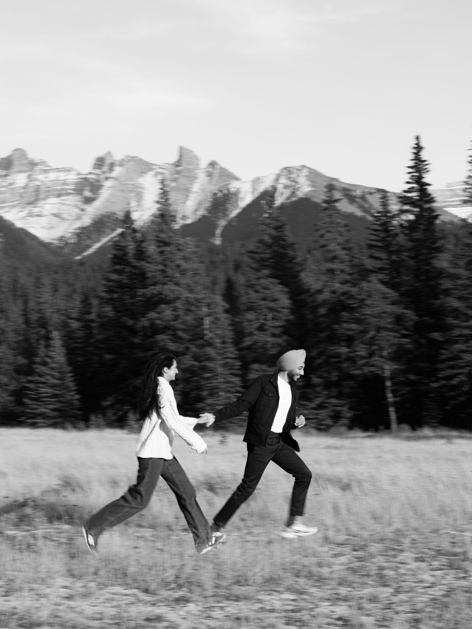 Black and white photo of a couple running joyfully through a mountain meadow with snowy peaks behind them.
