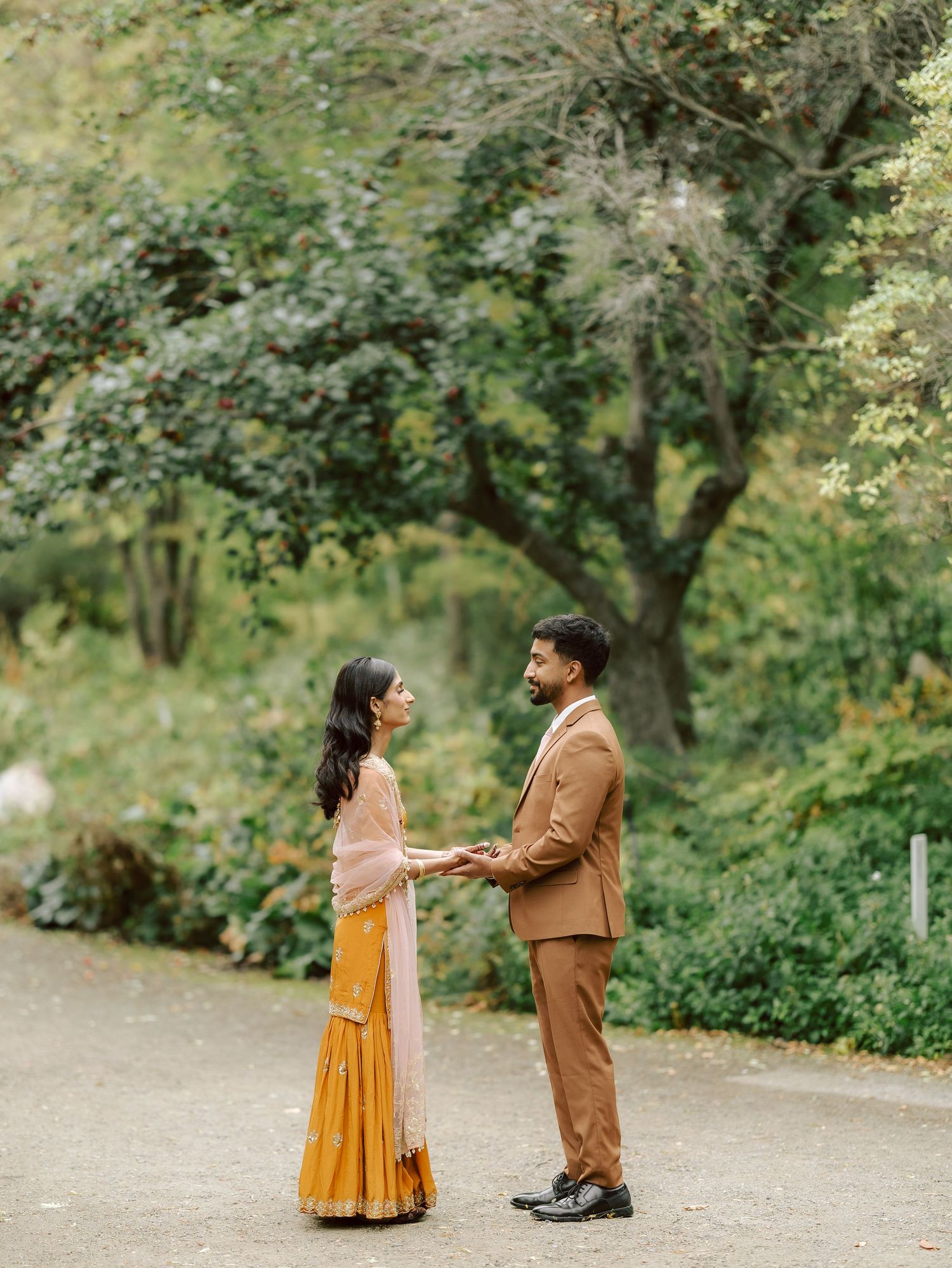South Asian wedding couple in traditional attire hold hands on garden path surrounded by greenery.