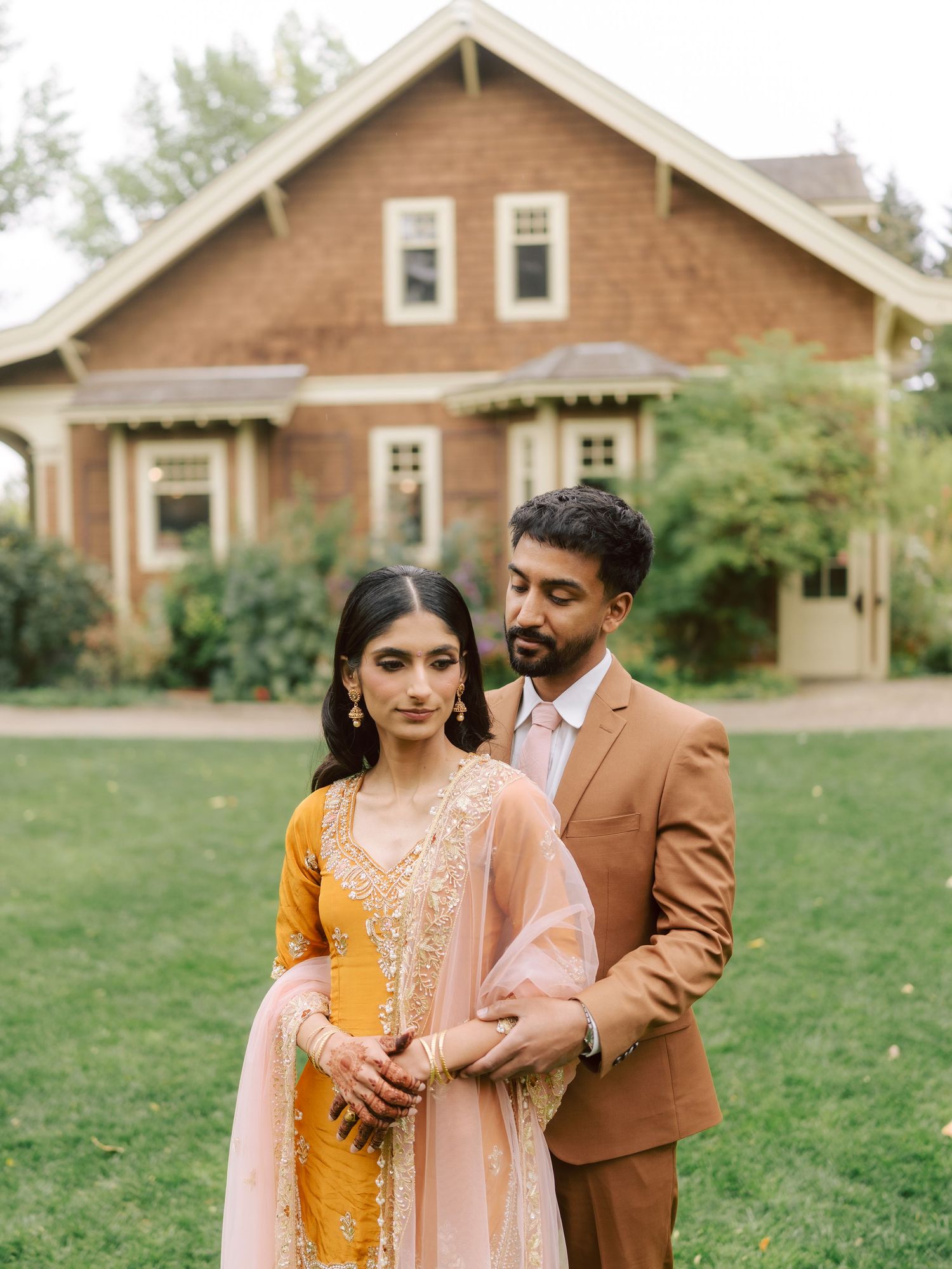 South Asian couple in traditional attire stands before rustic brick house.