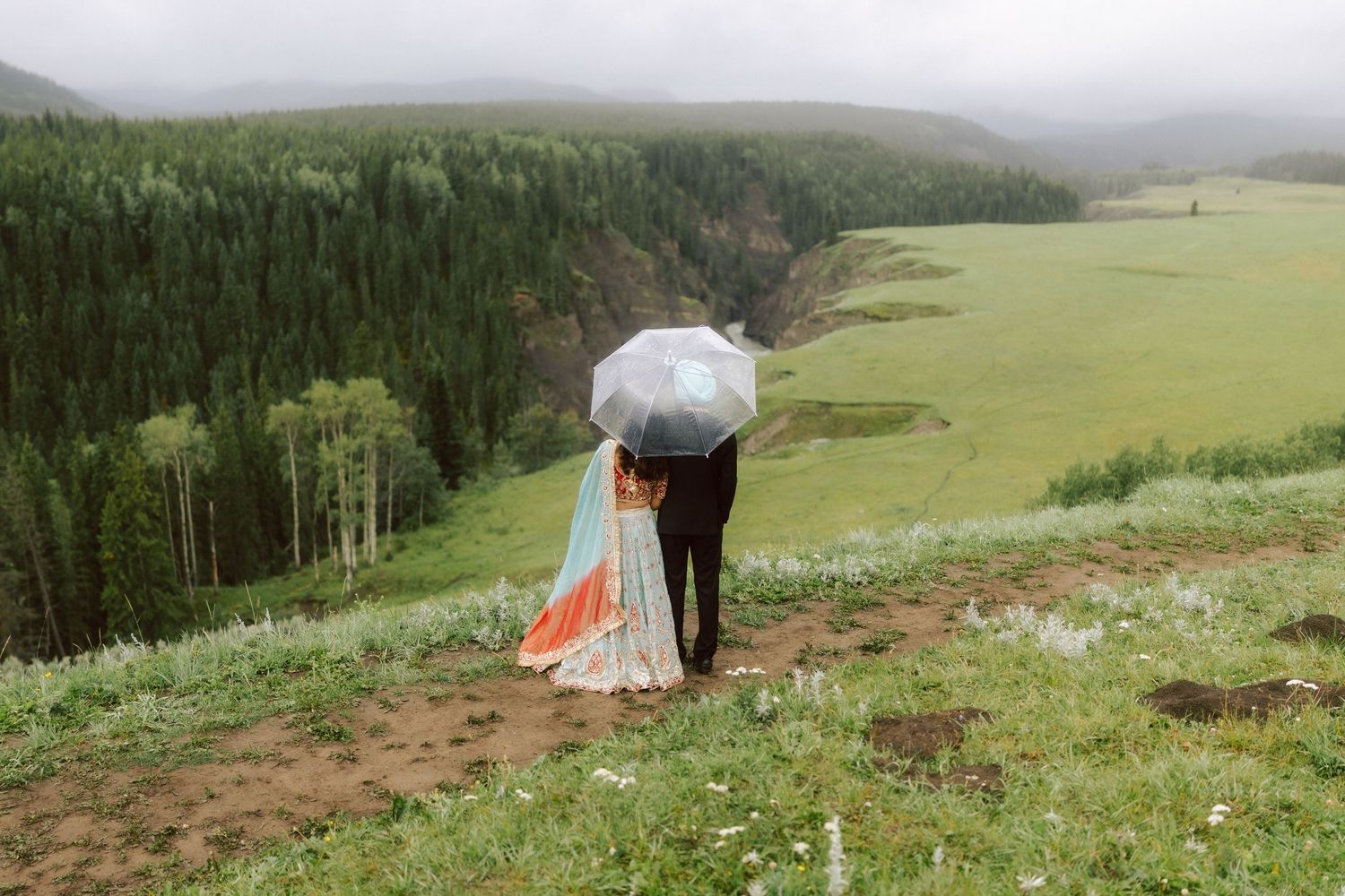 South Asian wedding couple sharing clear umbrella on rainy hilltop with green forest valley below.