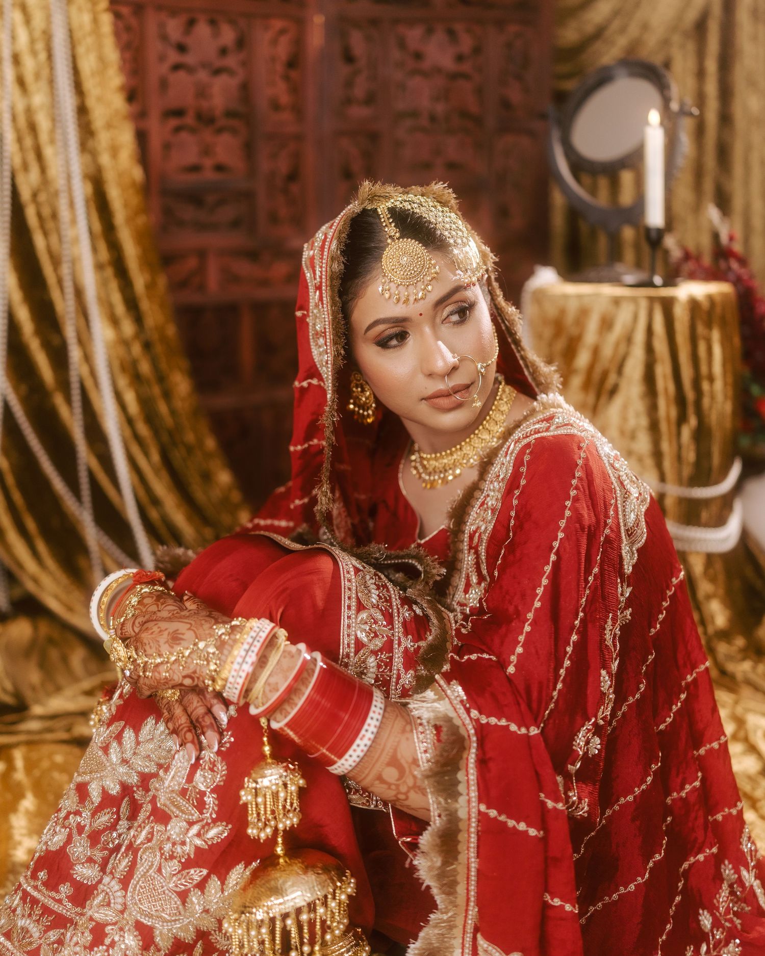 Indian bride in red embroidered lehenga with gold jewelry and maang tikka, posed in ornate golden setting.