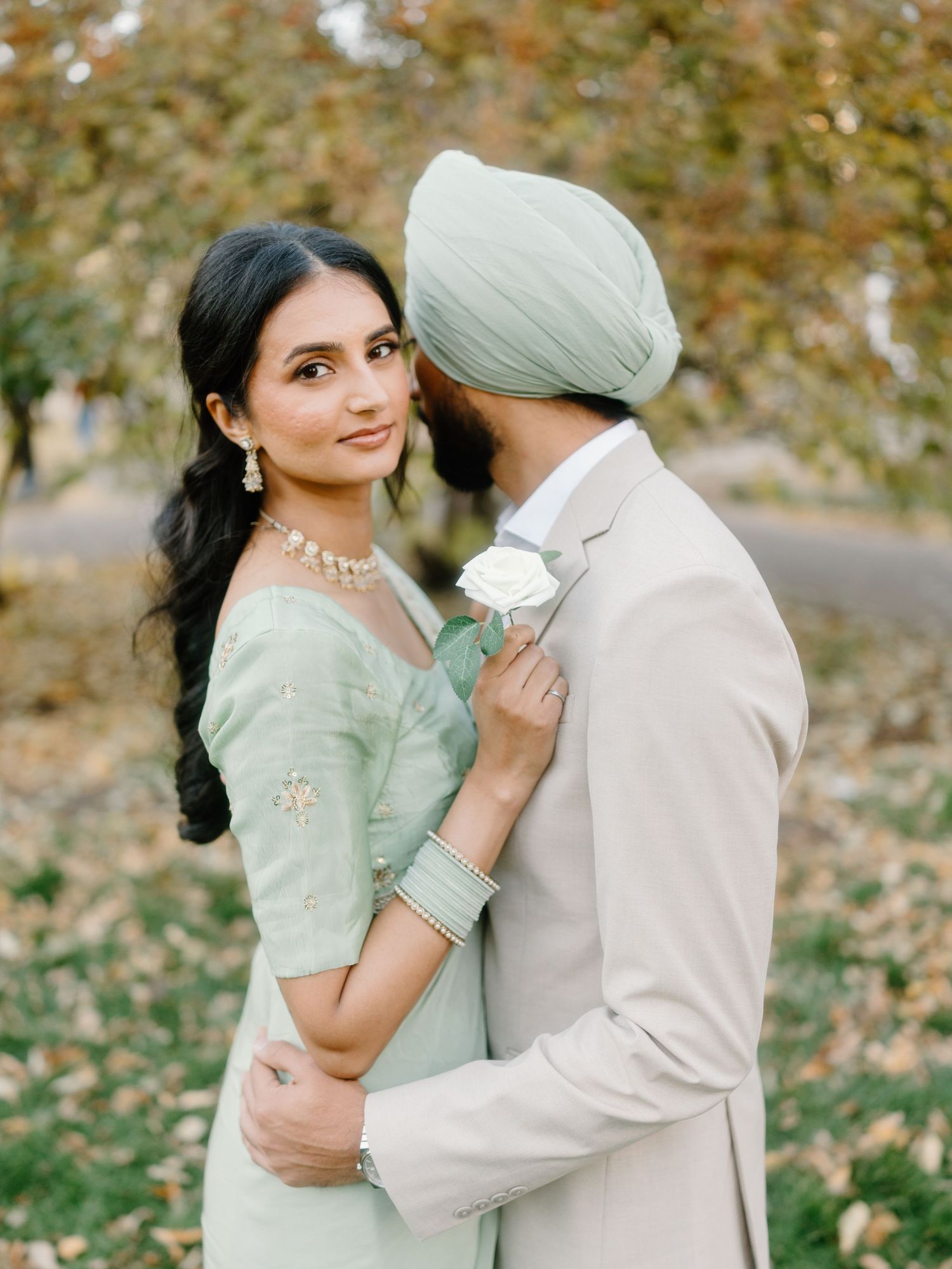 Sikh couple embracing tenderly outdoors, bride in mint saree and groom in beige suit with white rose.