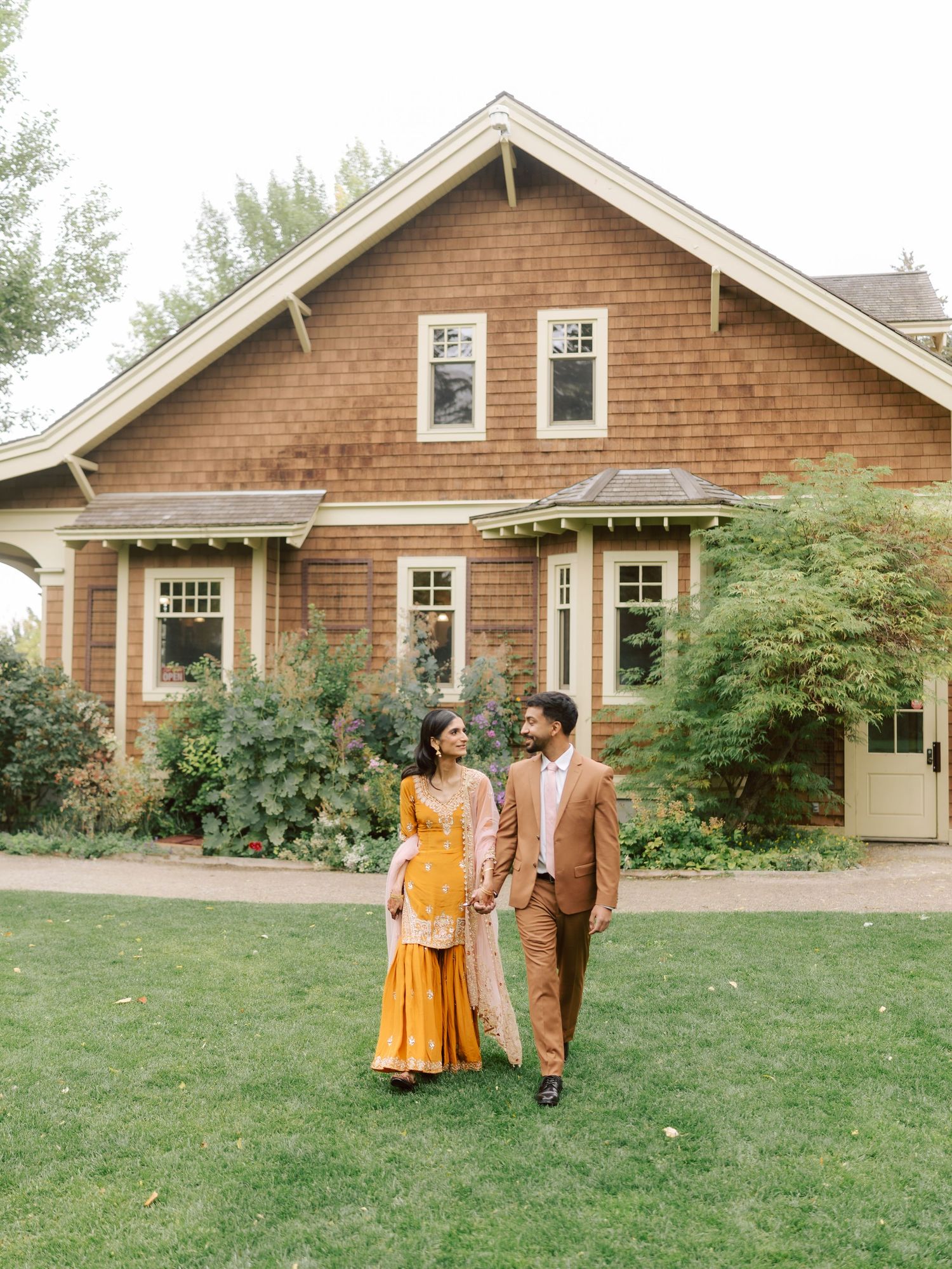 South Asian couple in traditional wedding attire walk hand-in-hand on green lawn before a craftsman home.