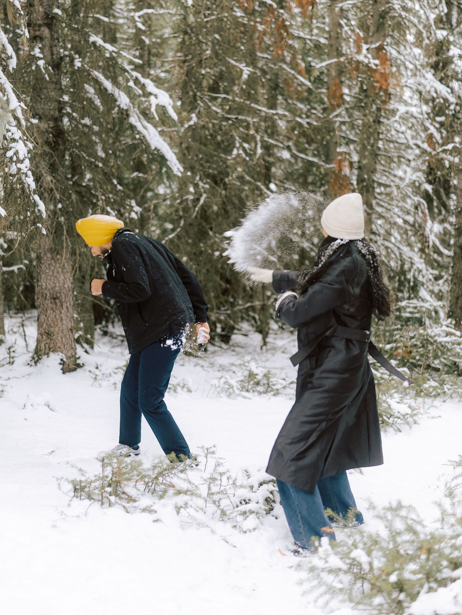 Couple throwing snowballs at each other in a snowy pine forest during a winter day.