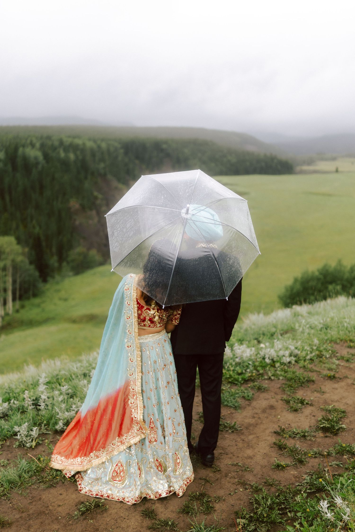 Indian bride and groom standing under transparent umbrella overlooking misty green valley on wedding day.