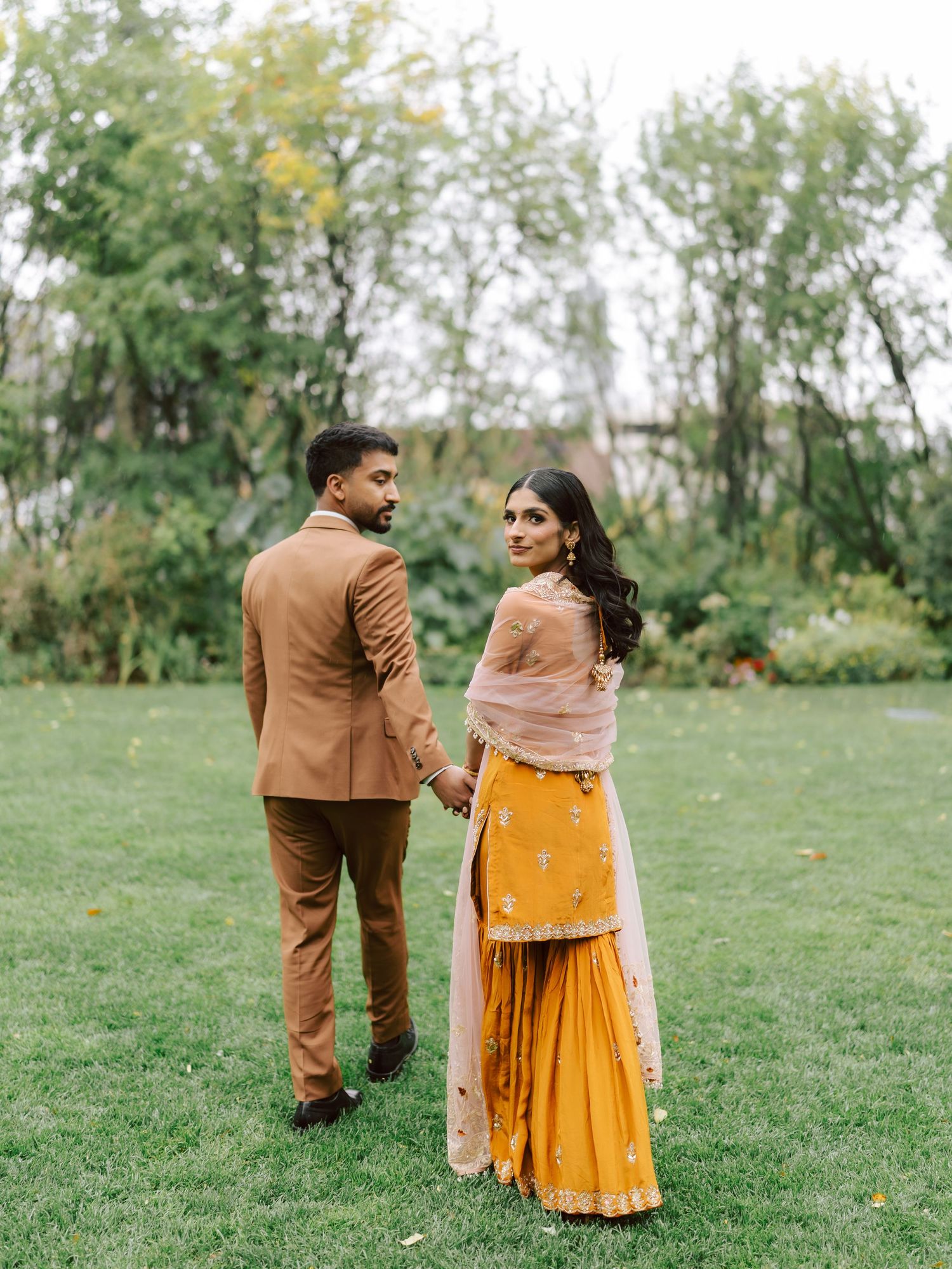 South Asian wedding couple holding hands look back at camera on lush green lawn.