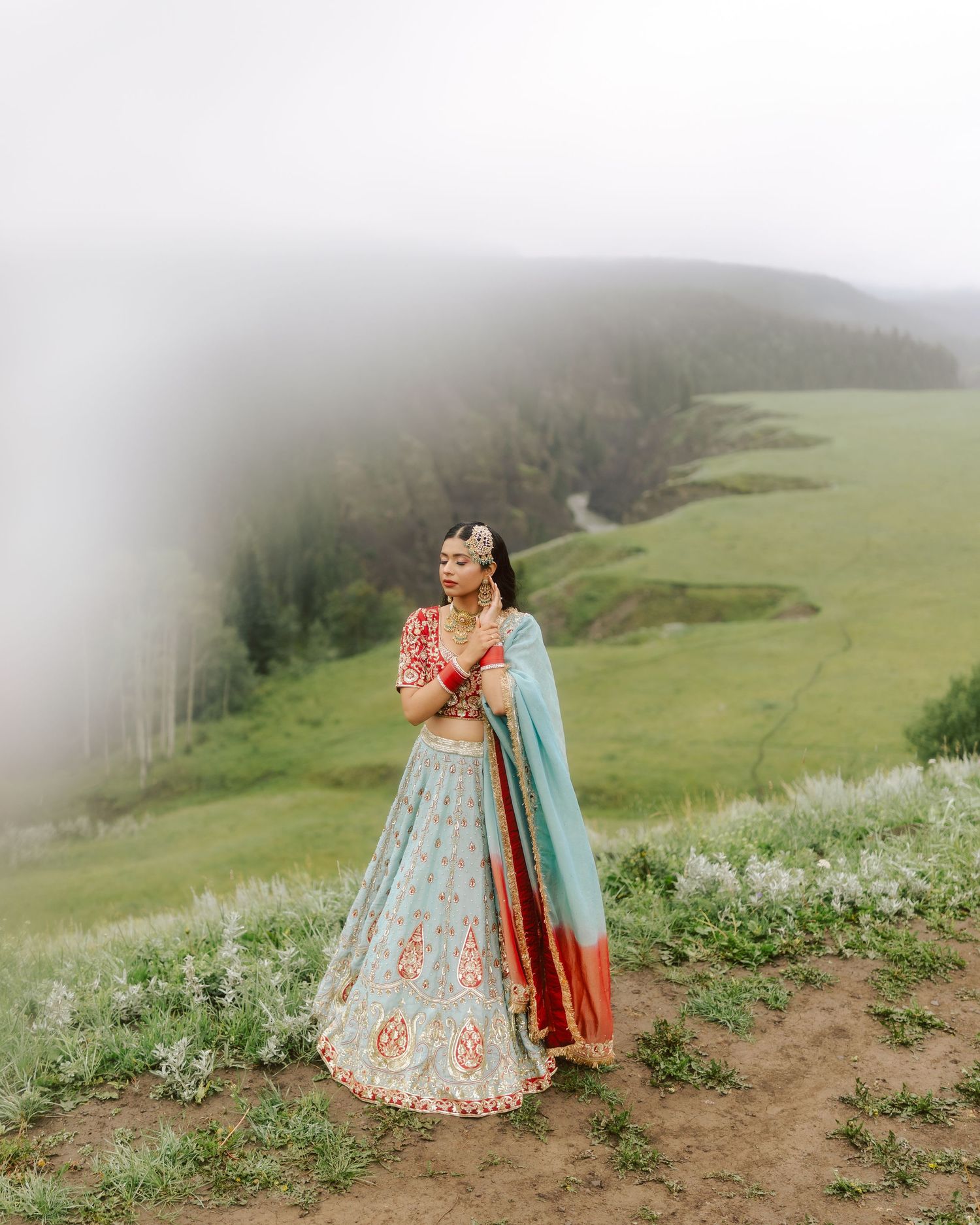 Indian bride in blue and red lehenga standing on misty hilltop with lush green valley below.