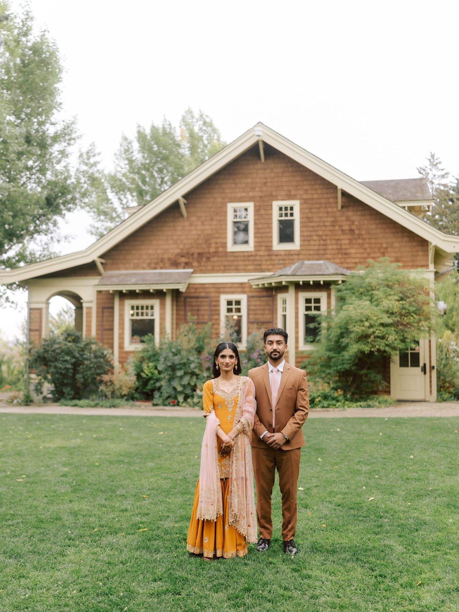 South Asian couple in traditional attire stand before a charming craftsman house on their wedding day.