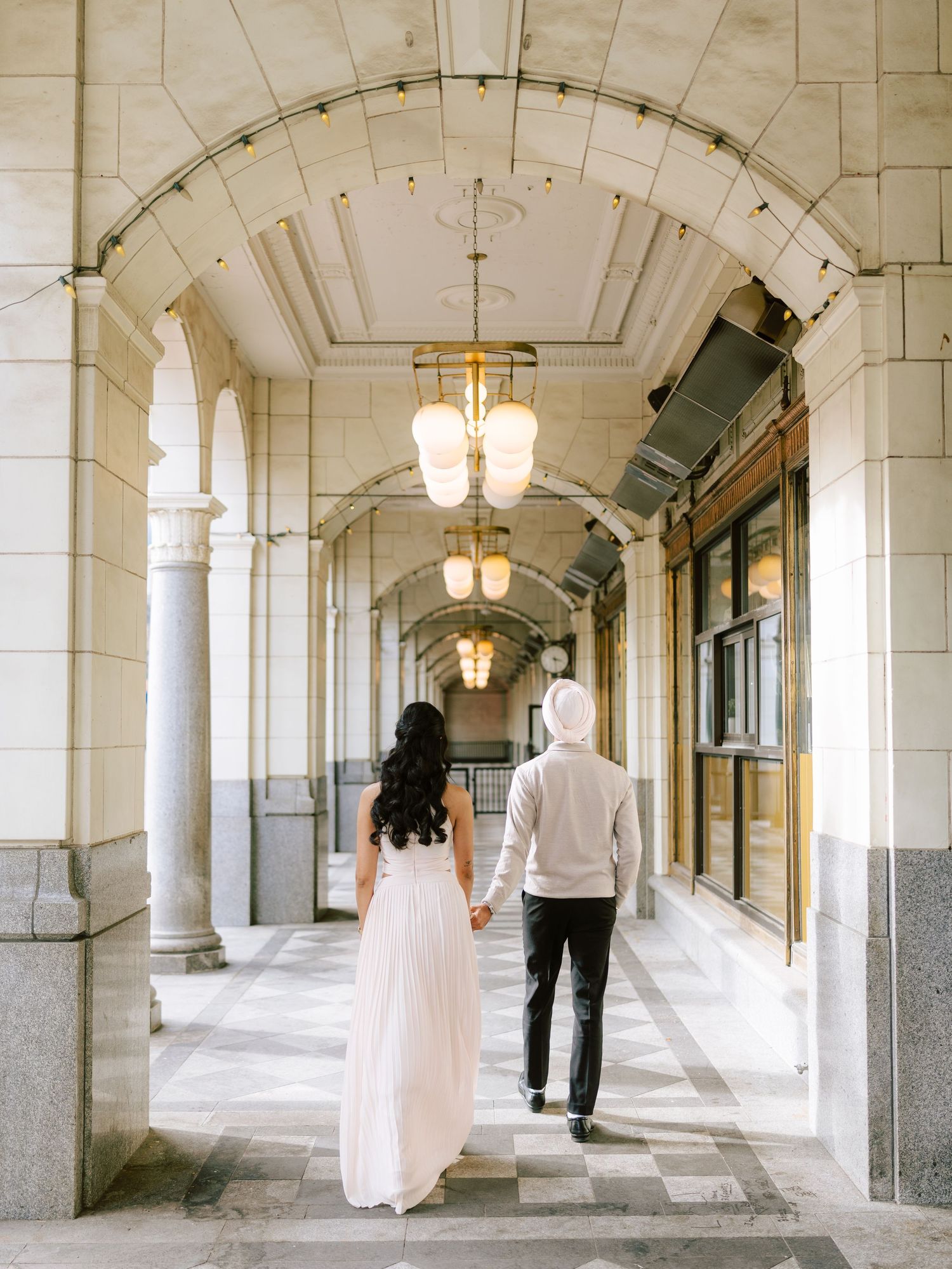 Couple walks hand in hand through an elegant arched hallway with ornate pendant lights.