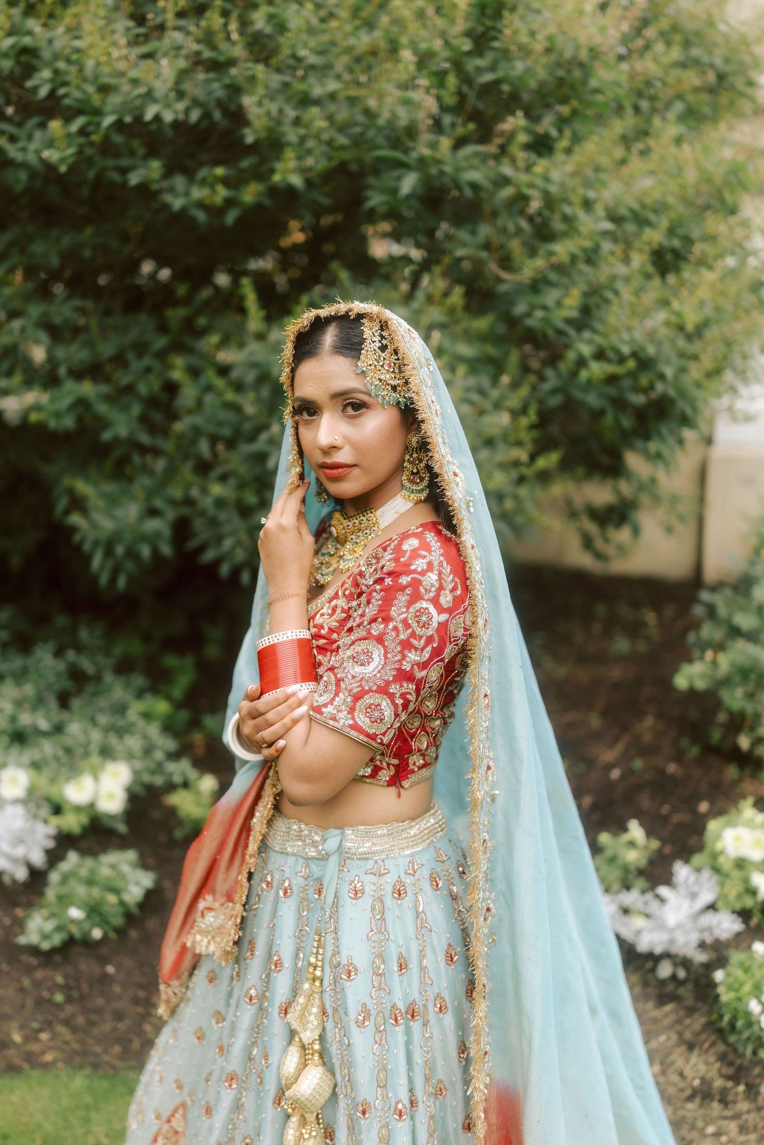 Indian bride in red and blue lehenga with gold jewelry and dupatta poses gracefully in a garden setting.