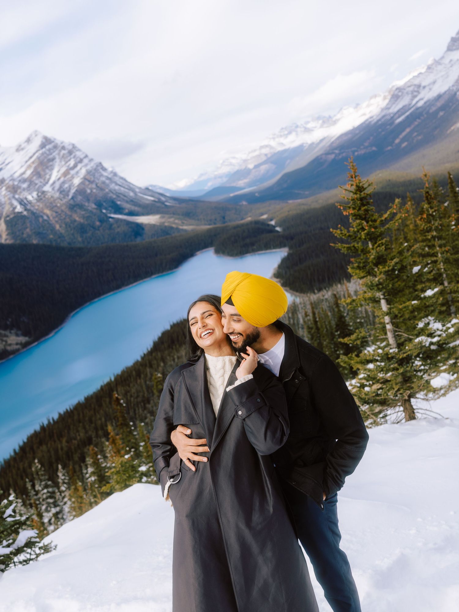 Couple laughing and embracing in snow with vivid blue Peyto Lake and mountains behind.