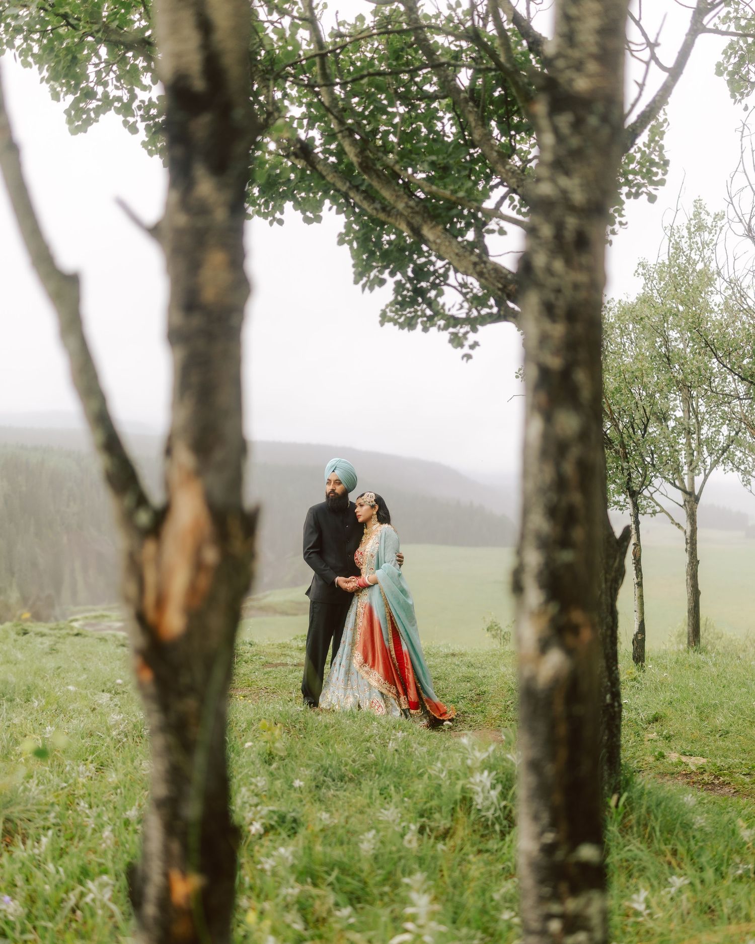 Sikh couple embracing in misty green meadow framed by birch trees during outdoor wedding photoshoot.