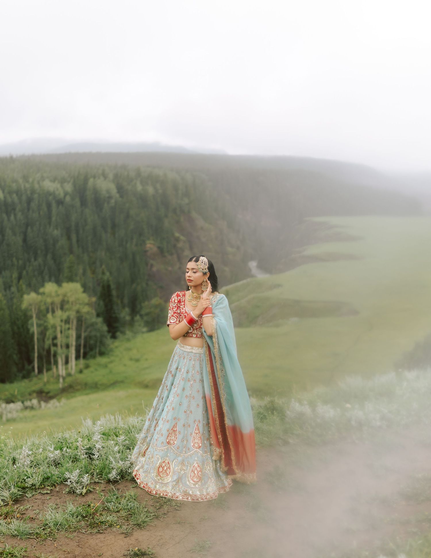 Sikh bride in blue and red lehenga standing on misty mountain cliff with forest valley below.