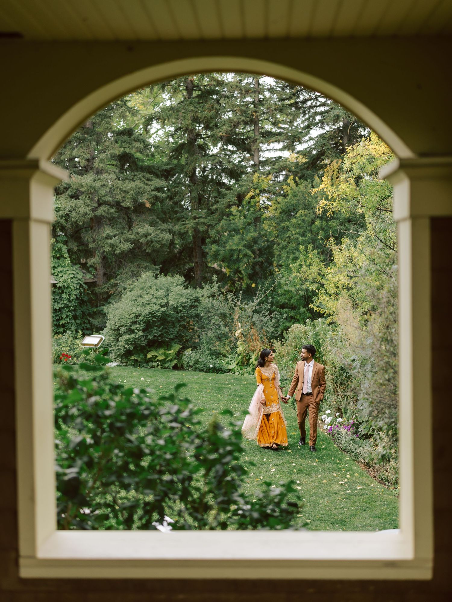 Couple in vibrant orange dress and brown suit walking hand-in-hand through lush garden, framed by an arched window.