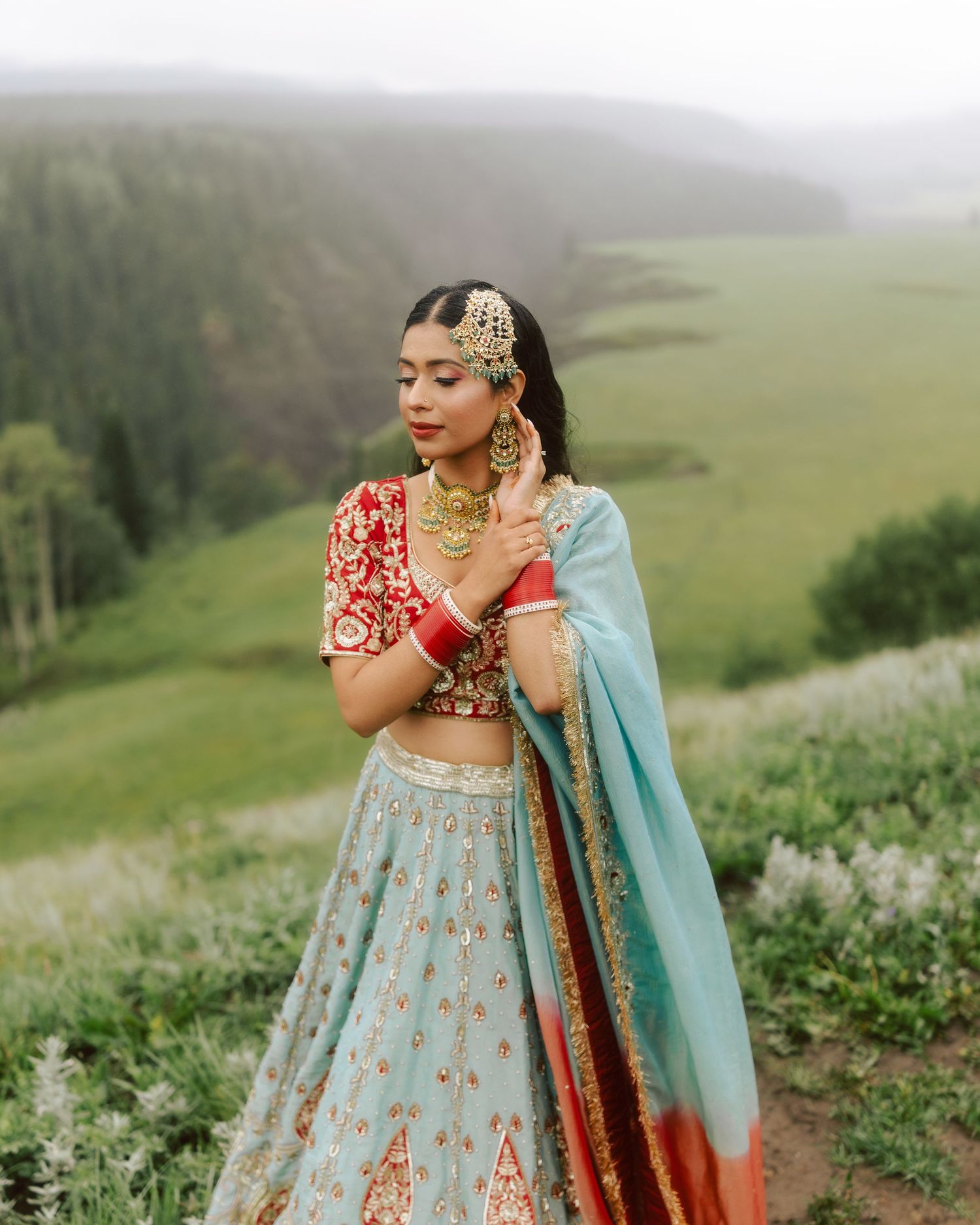 Sikh bride in red and blue lehenga with gold jewelry posing on lush green misty hillside.