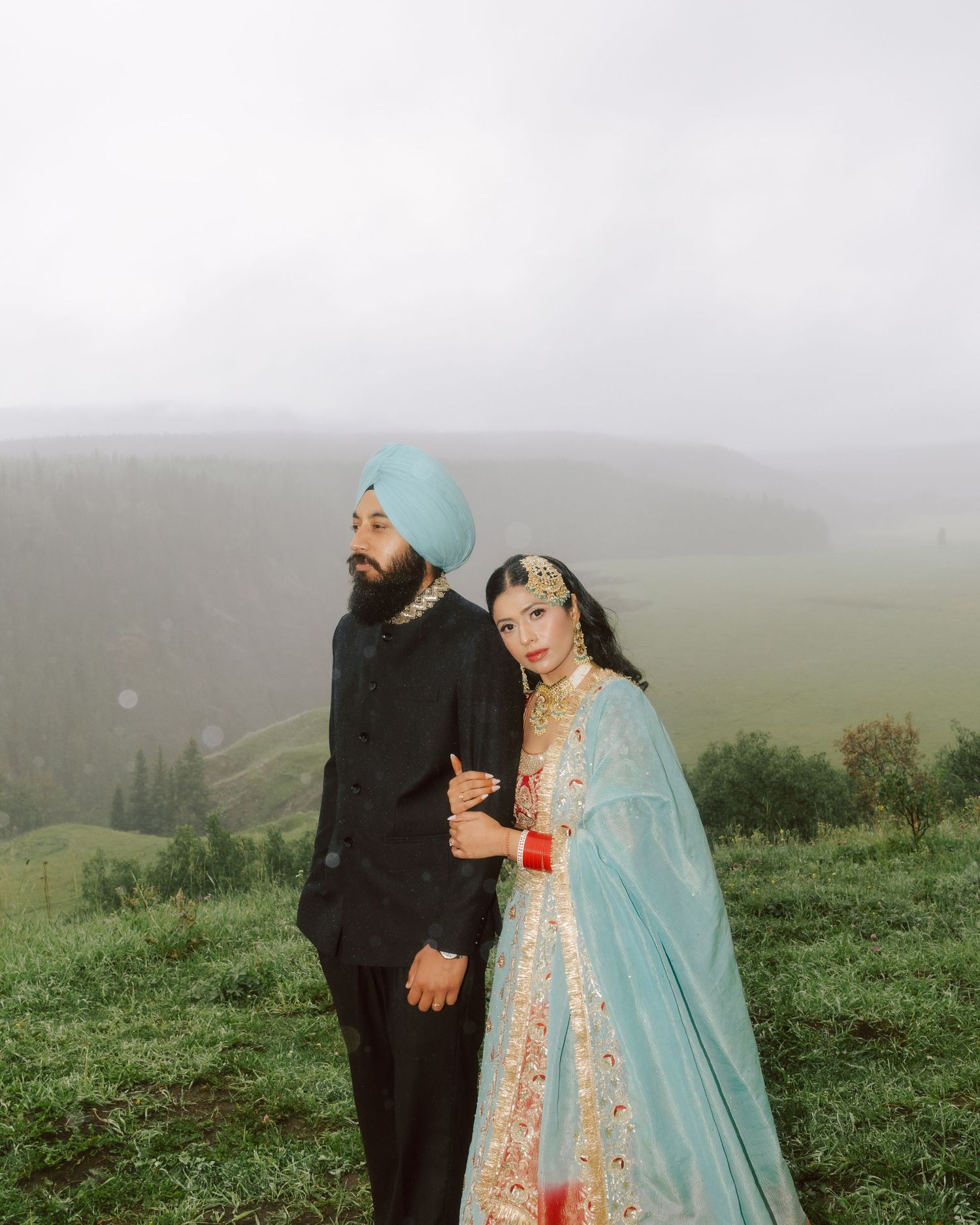 Sikh couple in traditional wedding attire poses on misty green hillside.