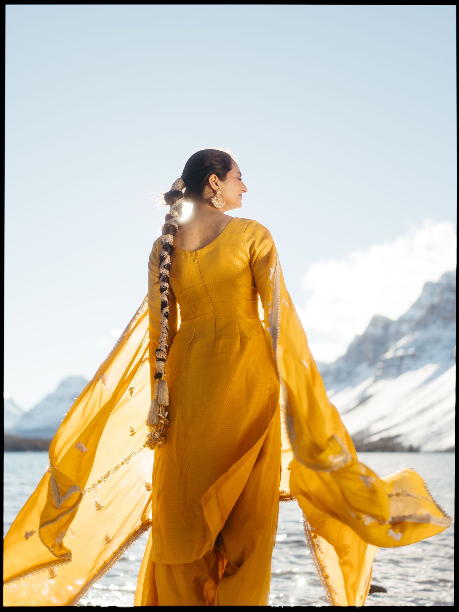 Woman in golden yellow anarkali suit with dupatta billowing in wind by snowy mountain lake.