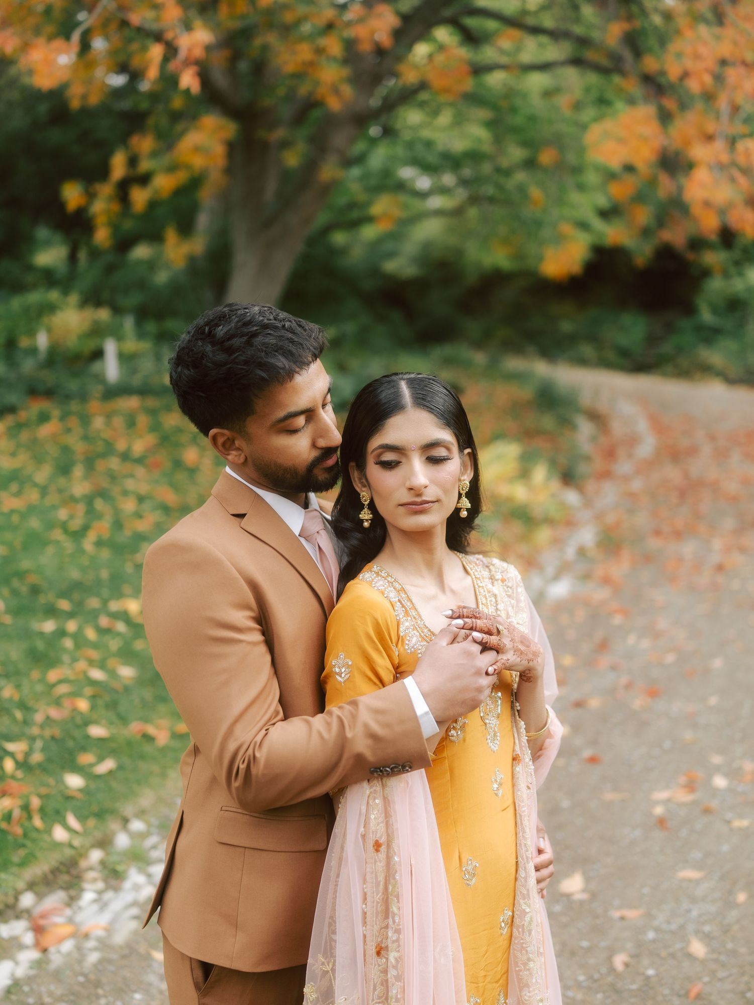 South Asian couple embracing on autumn path, groom in tan suit and bride in yellow embroidered lehenga.