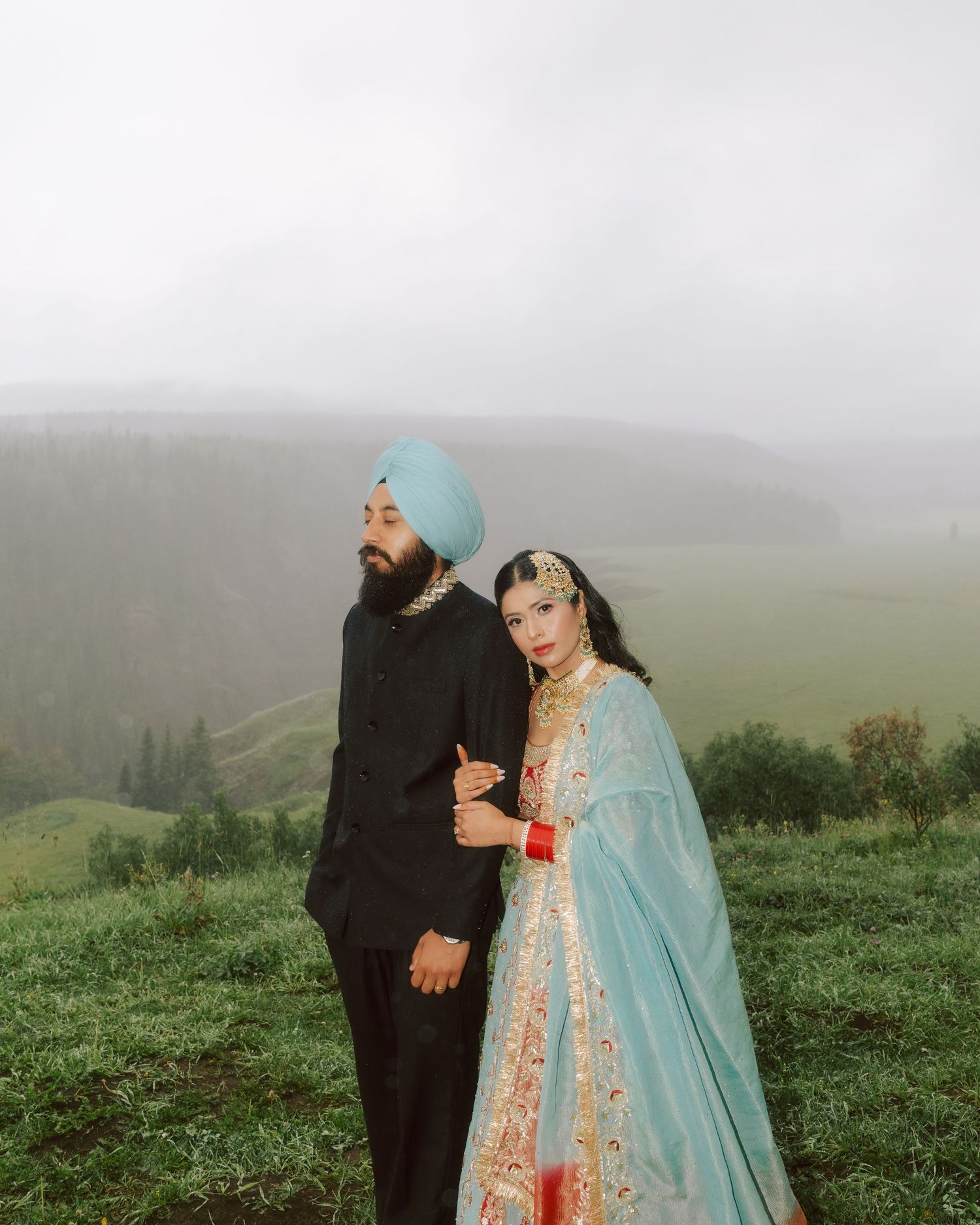 Sikh wedding couple in traditional attire standing on foggy green hillside with valley views.