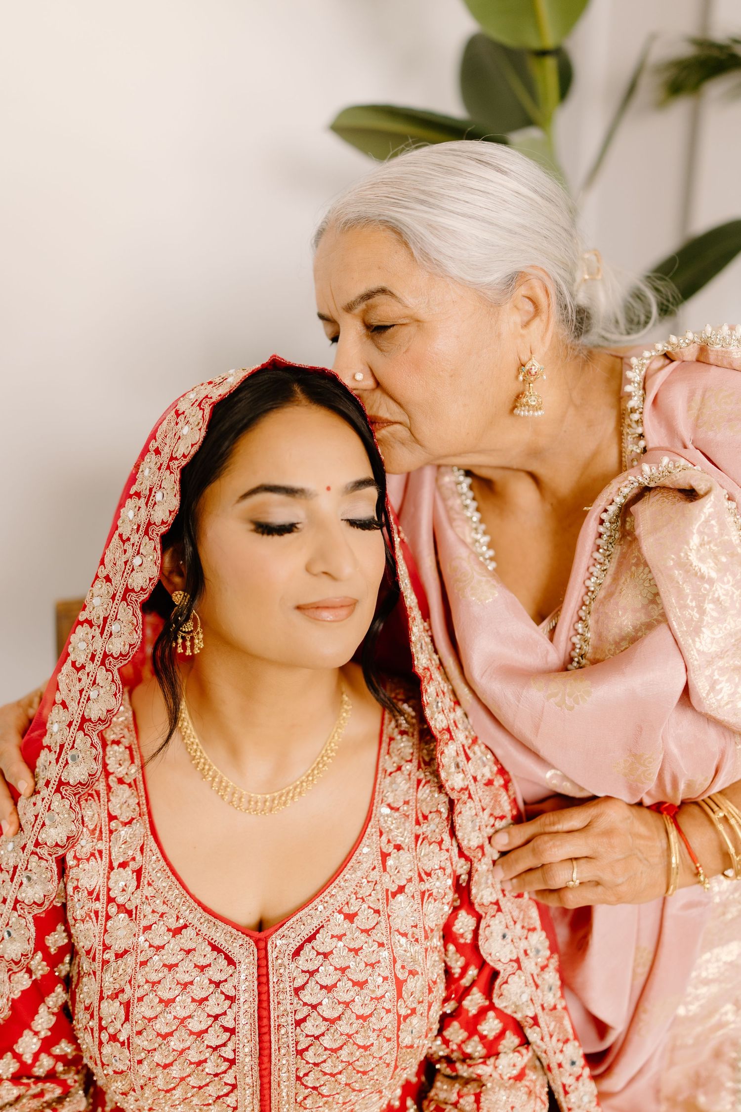Elderly woman in pink sari kisses bride in red lehenga on forehead during Indian wedding ceremony.