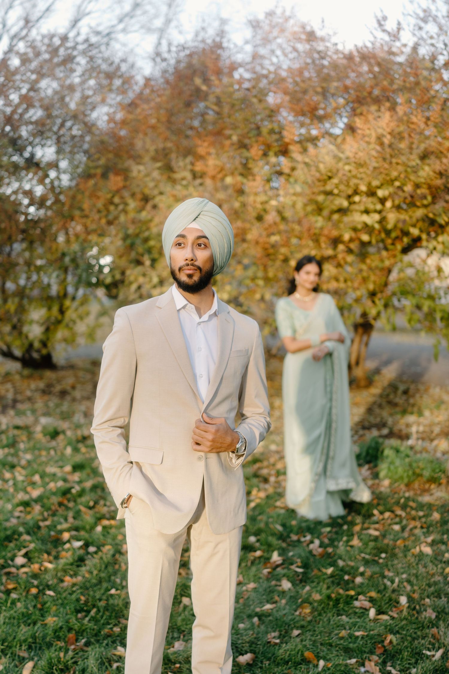 Sikh groom in beige suit and sage turban stands confidently outdoors with bride in mint saree behind.