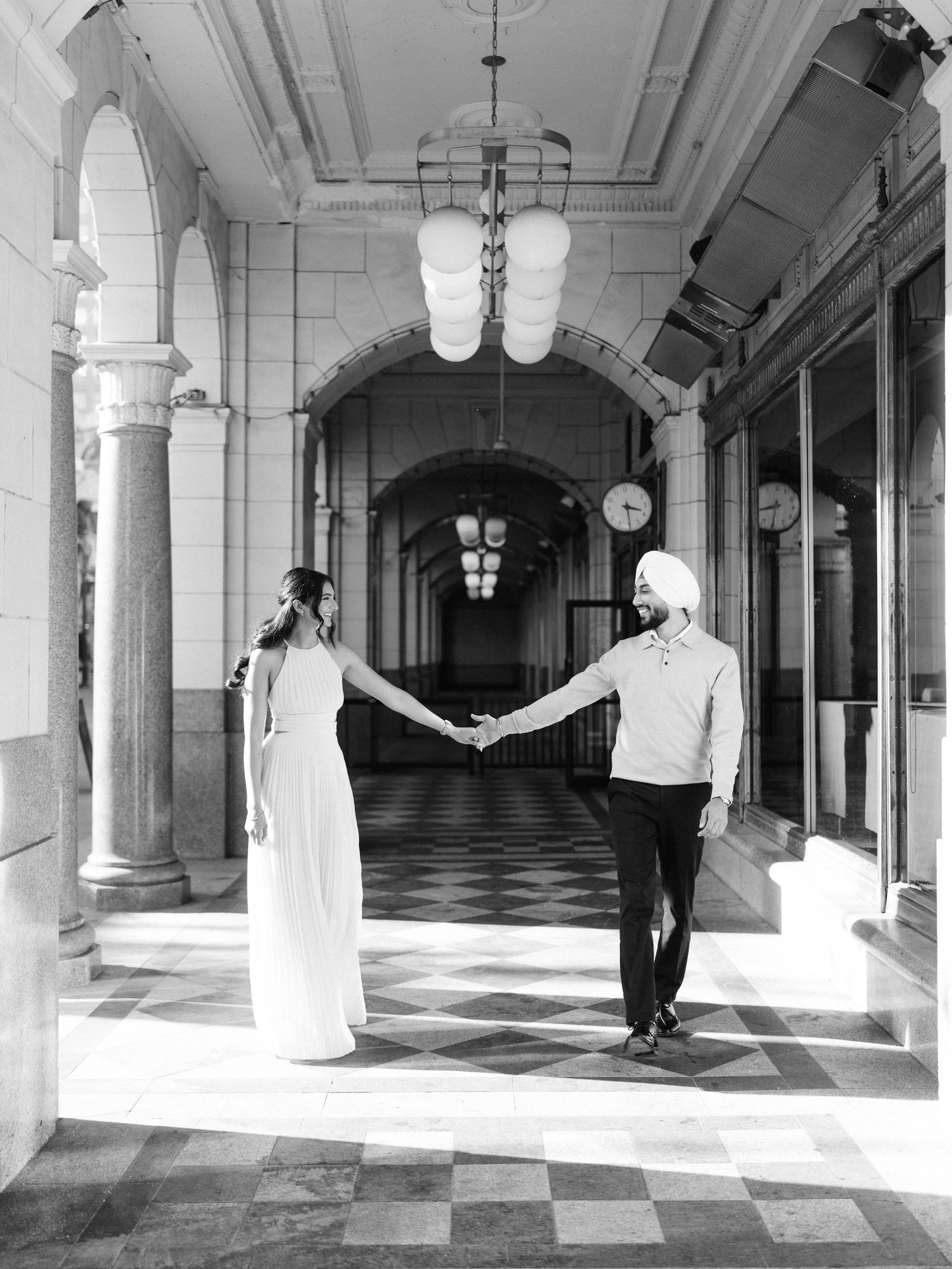 Couple holds hands in a grand black and white hallway with ornate columns and checkered floors.
