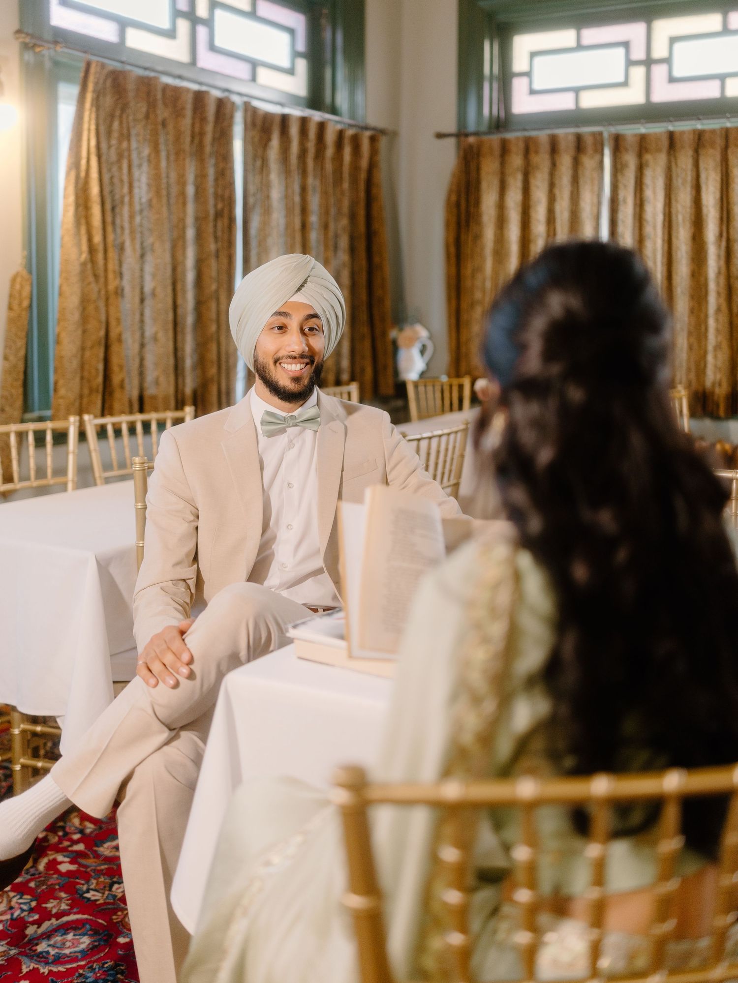 Handsome Sikh groom in beige suit and sage turban smiling at bride during wedding ceremony.