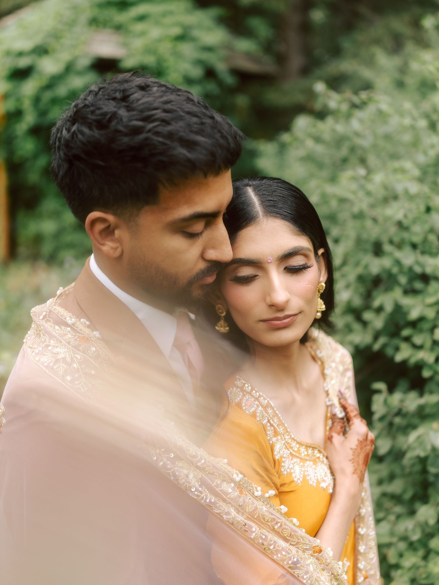 South Asian groom kisses brides temple as she leans into him in sunlit garden setting.