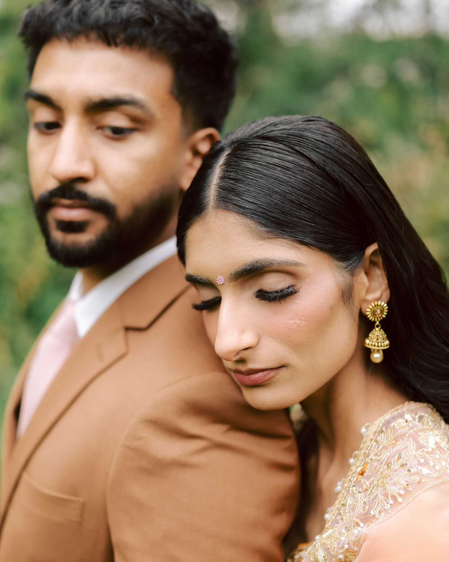 Indian bride in red bridal attire smiling as groom adjusts her gold chandelier earring outdoors.