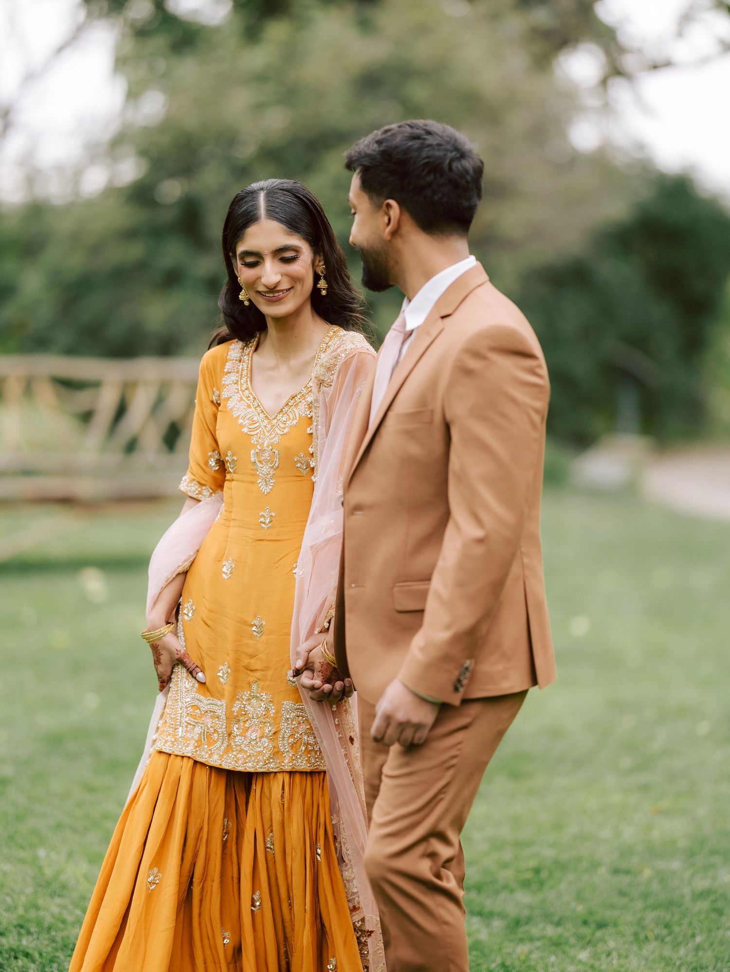 Groom in tan suit gazes at bride wearing yellow embroidered sharara with blush dupatta outdoors.