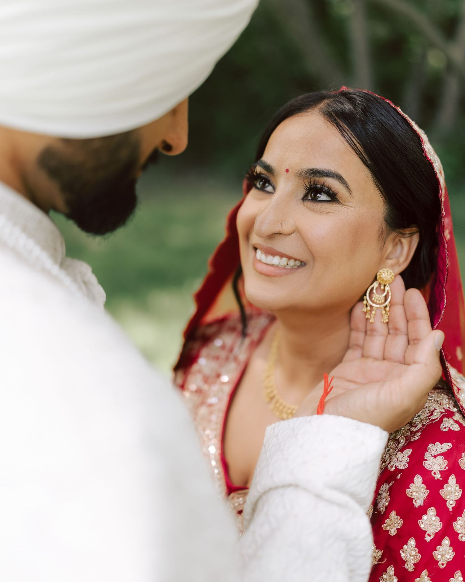 Sikh bride in red lehenga smiling radiantly as groom in white turban adjusts her gold earring outdoors.