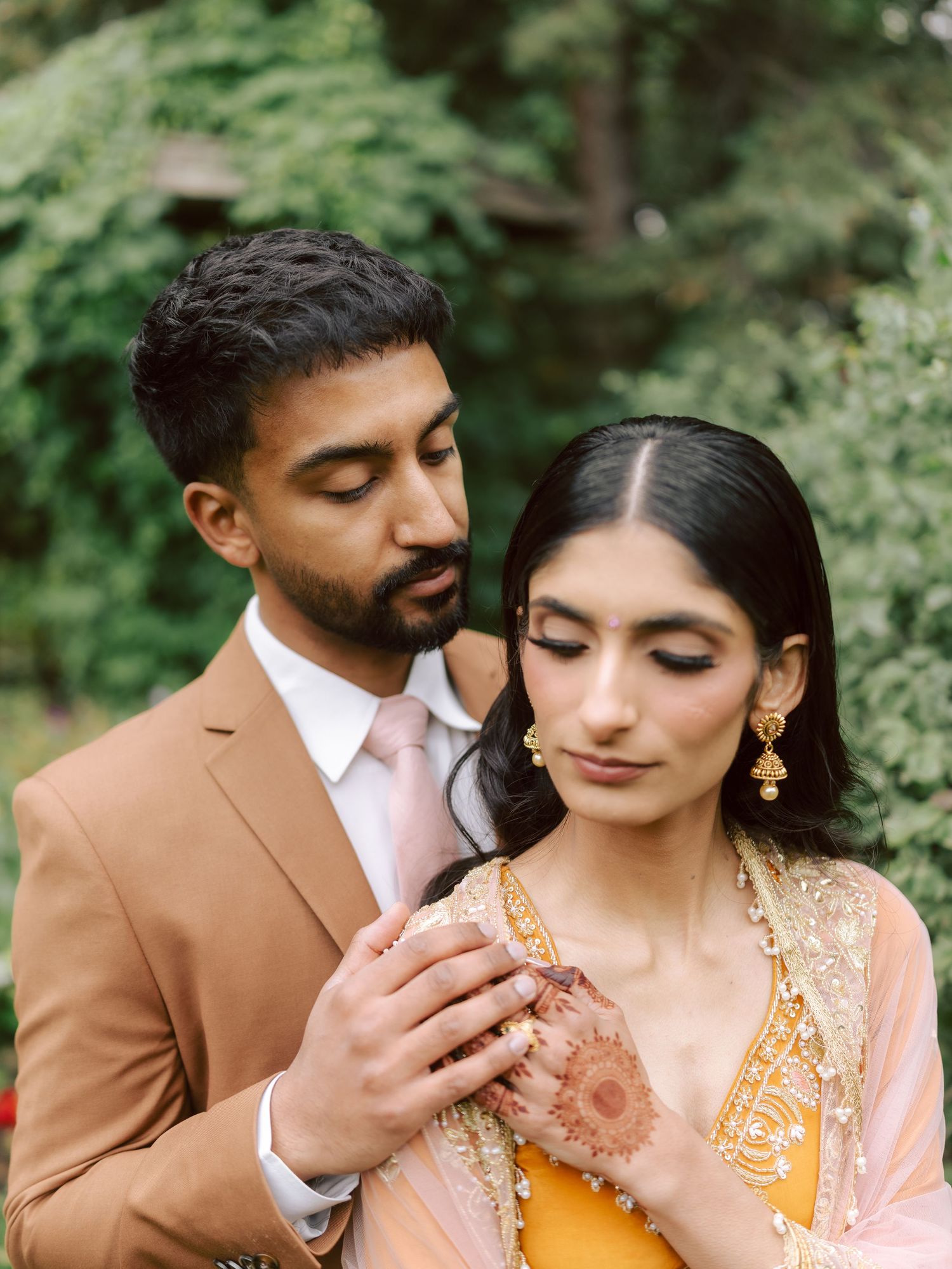 South Asian groom in caramel suit tenderly holds brides henna-adorned hand in garden setting.