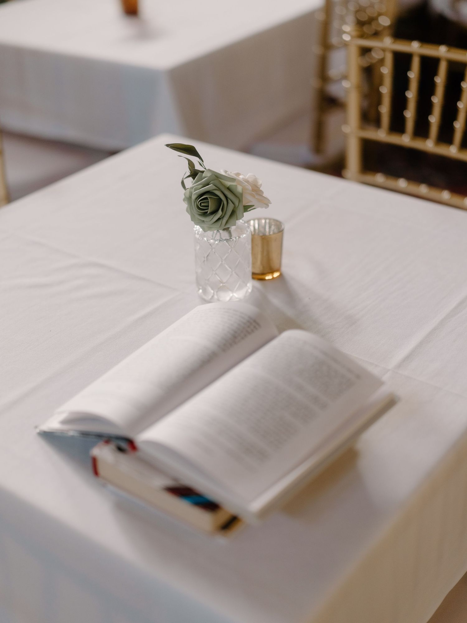 Open book with sage green rose and gold candle on white linen table at wedding venue.