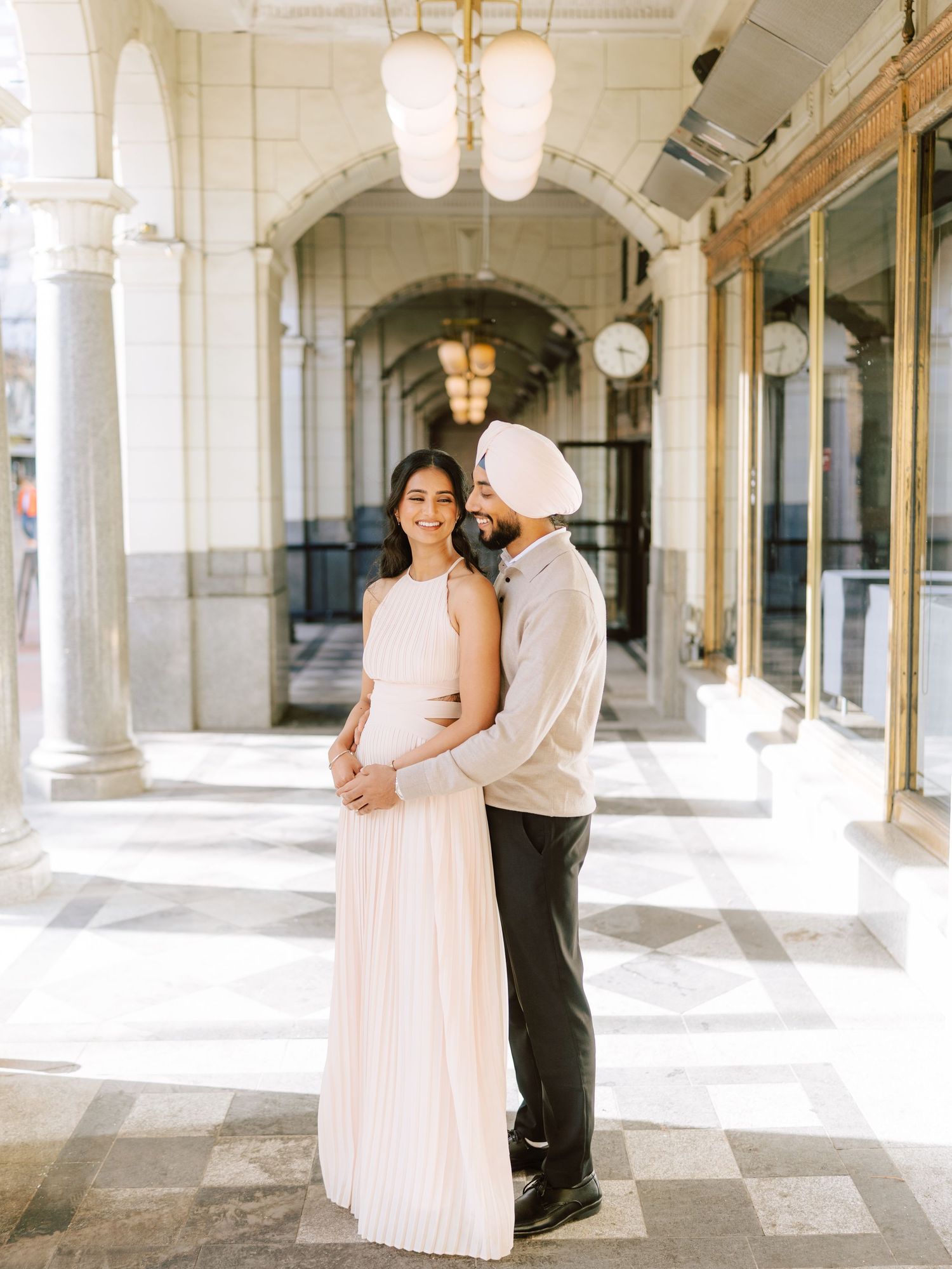 Smiling Sikh couple posing in sunlit classical arcade with checkered floors and chandeliers.